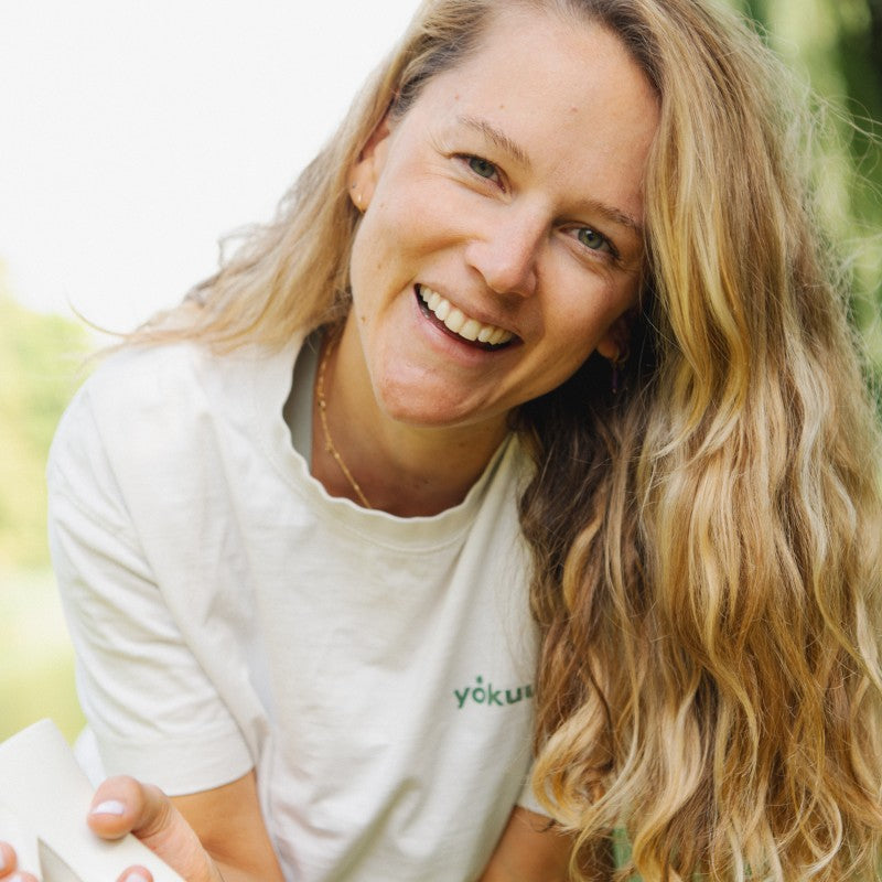 Woman with long blonde hair wearing a white shirt with a visible brand logo, sitting outdoors.